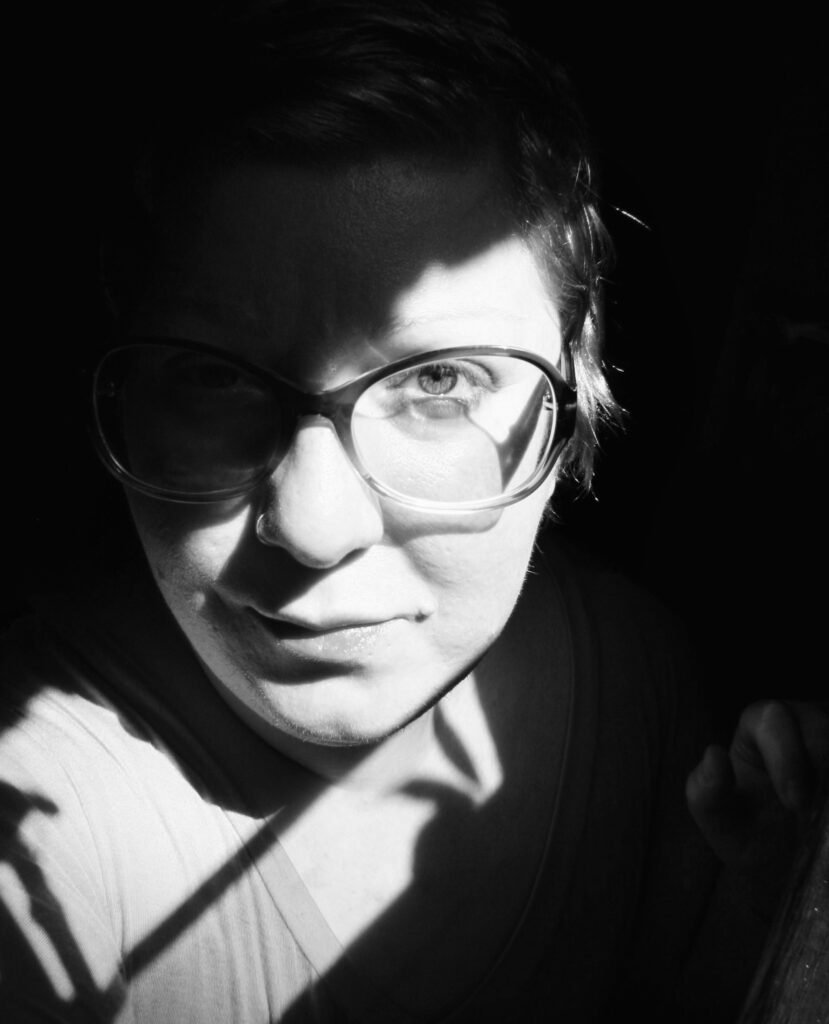 A black and white image of a human with short hair, wearing big retro reading glasses. They are looking directly at the camera lens with a neutral expression and partially in shadow. The light is illuminating the human's face in a diagonal direction coming from the top right of the image. 